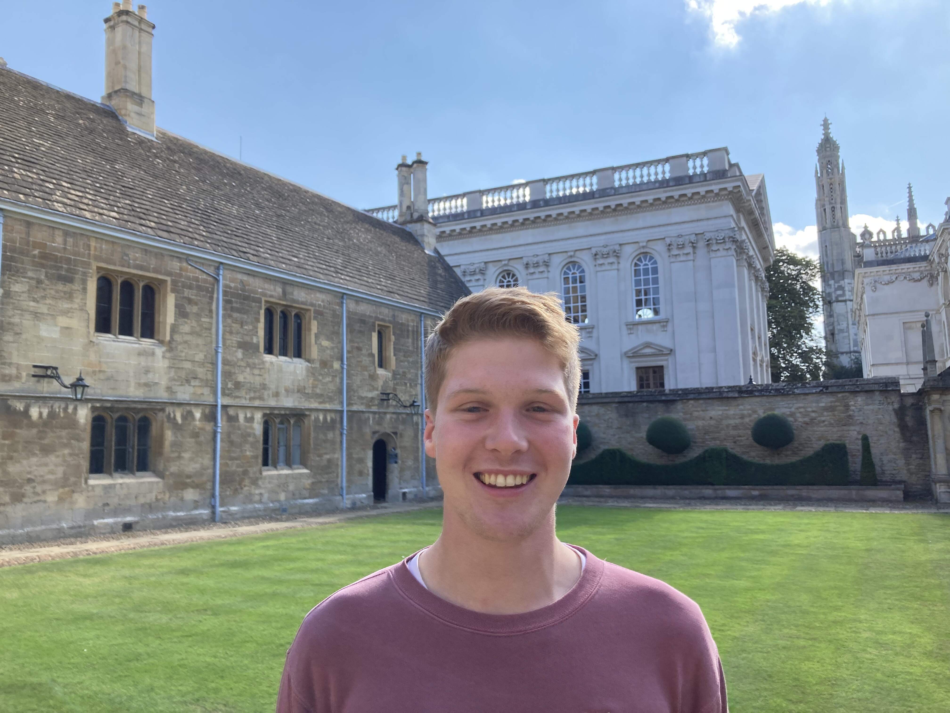 Picture of David smiling in front of Caius Court, wearing a red jumper, with Senate house in the background