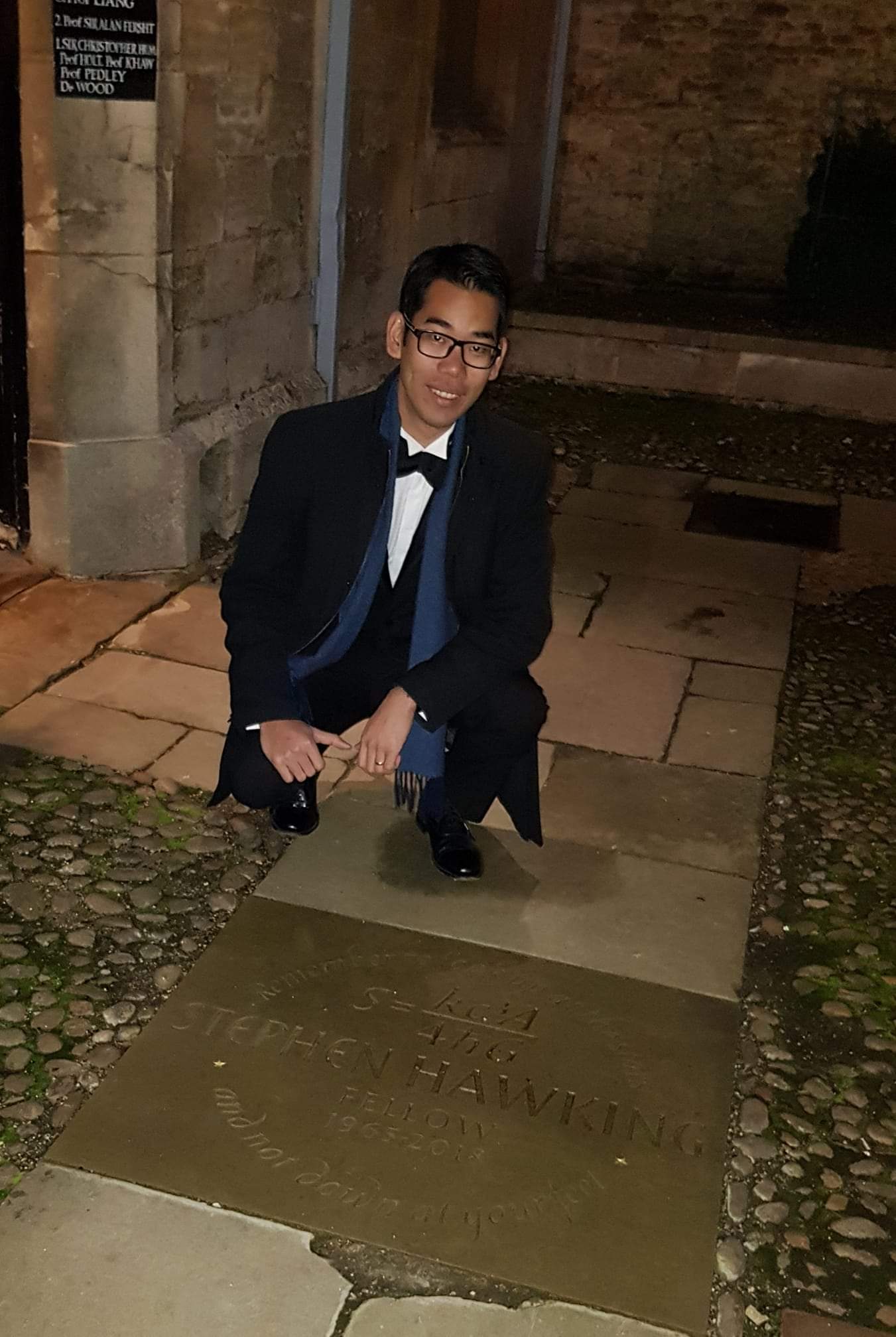 A man in a dinner jacket crouching next to a stone bearing the name of Professor Stephen Hawking