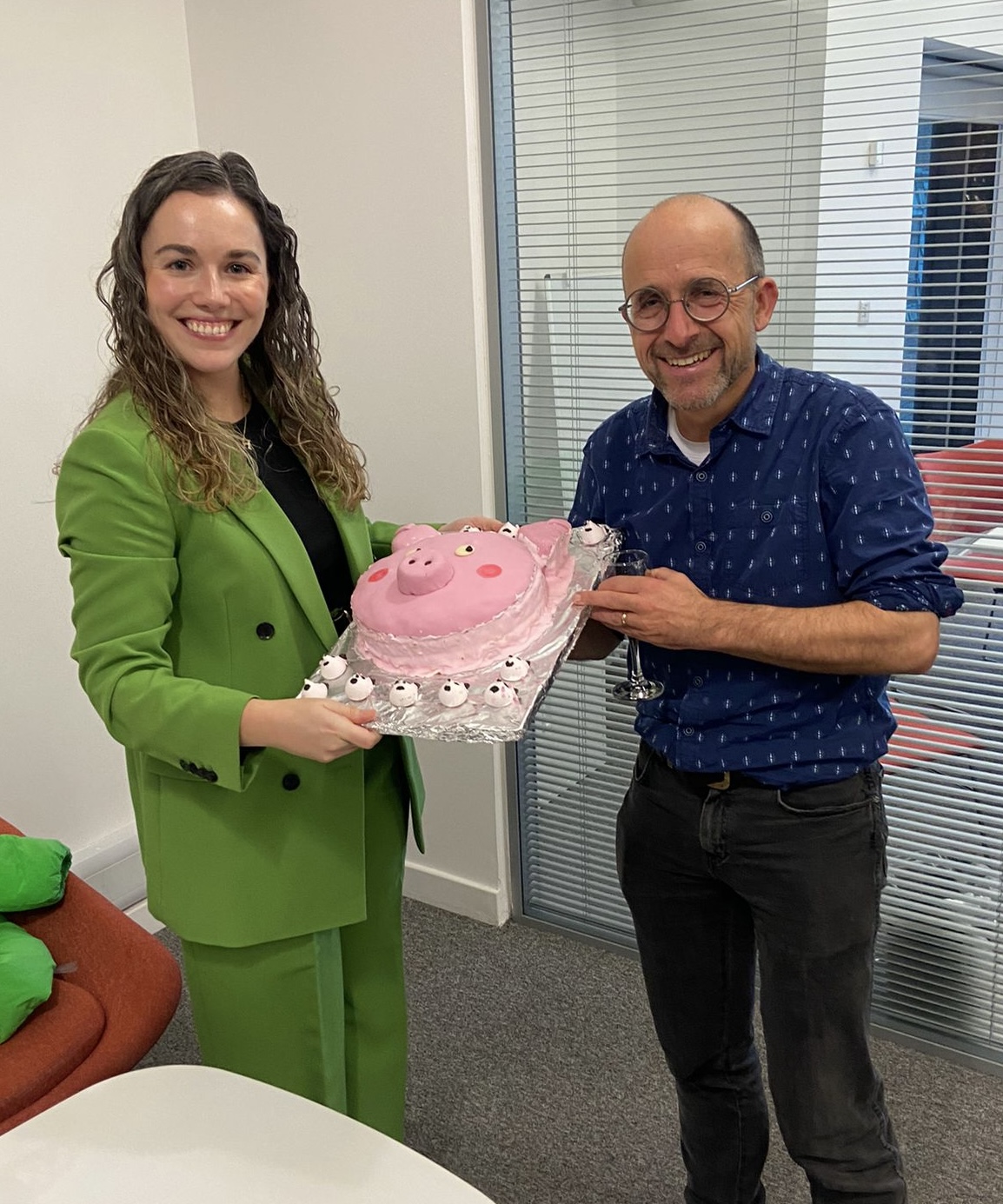 A woman in a green trouser suit and a man in a navy blue shirt with a cake shaped in the face of a pig