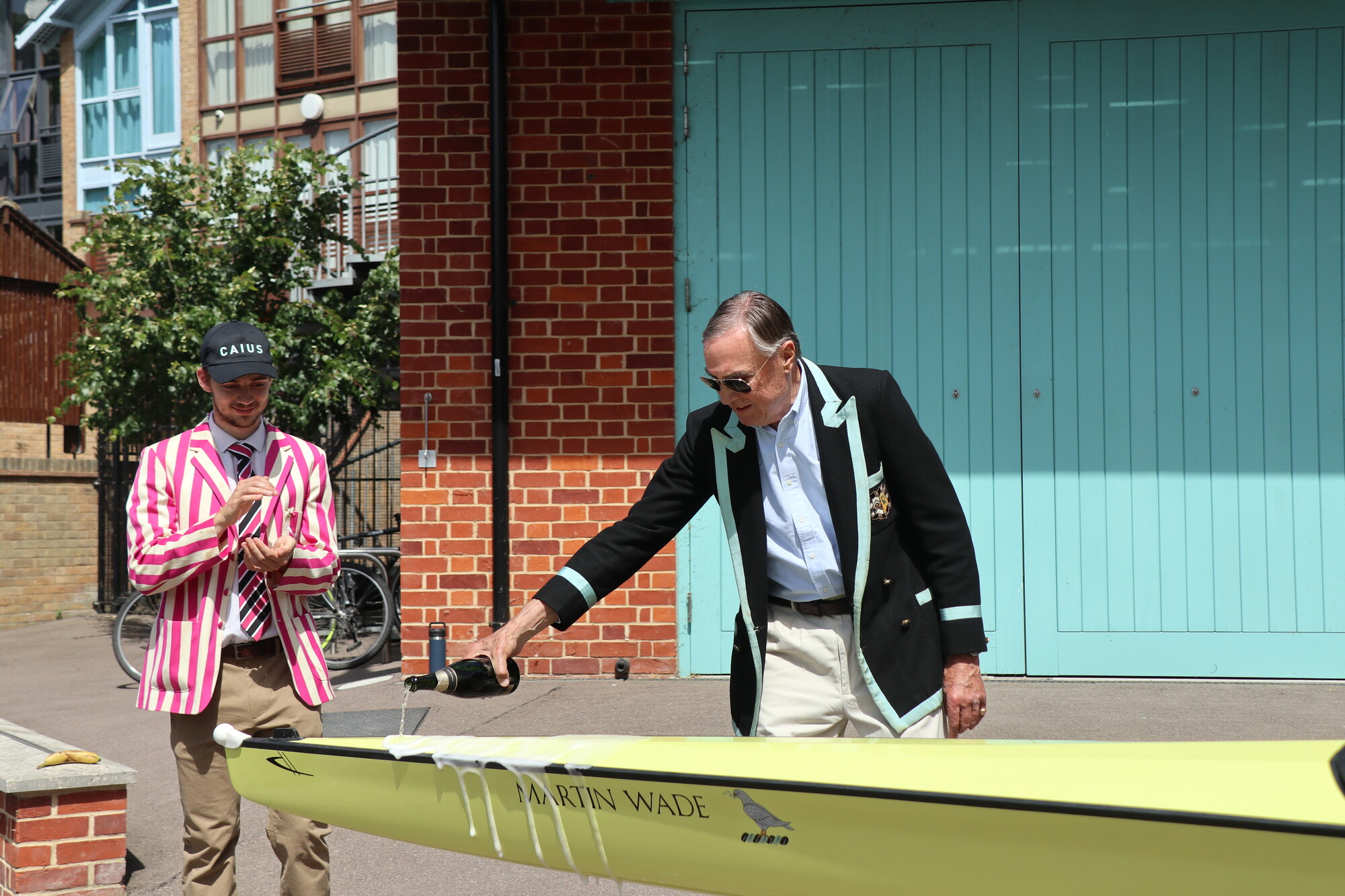 A man in a black blazer pours champagne over a rowing boat in a naming ceremony
