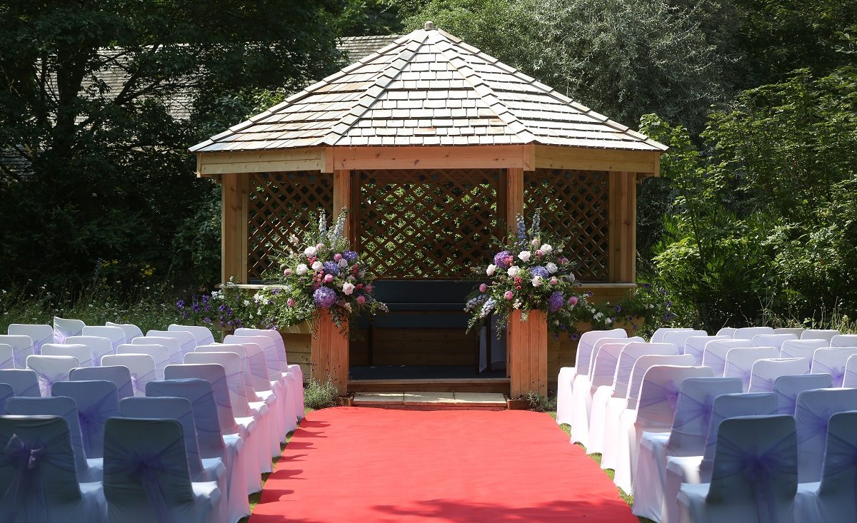 A cedar-wood wedding gazebo set for an outdoor wedding ceremony with a red carpet and chairs covered with white satin