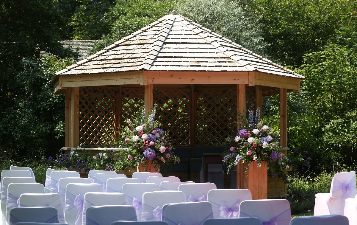 A cedar-wood gazebo set for an outdoor wedding ceremony with beautiful floral pedestals 