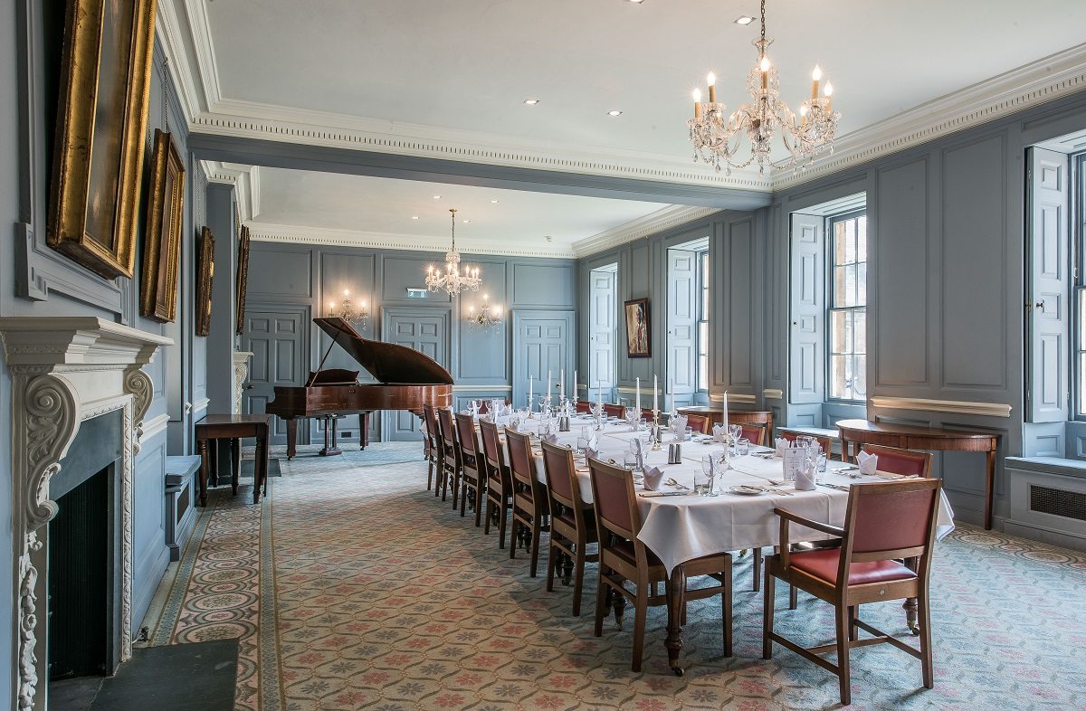 A large table set for dinner with a grand piano in the corner of the room and chandeliers lit